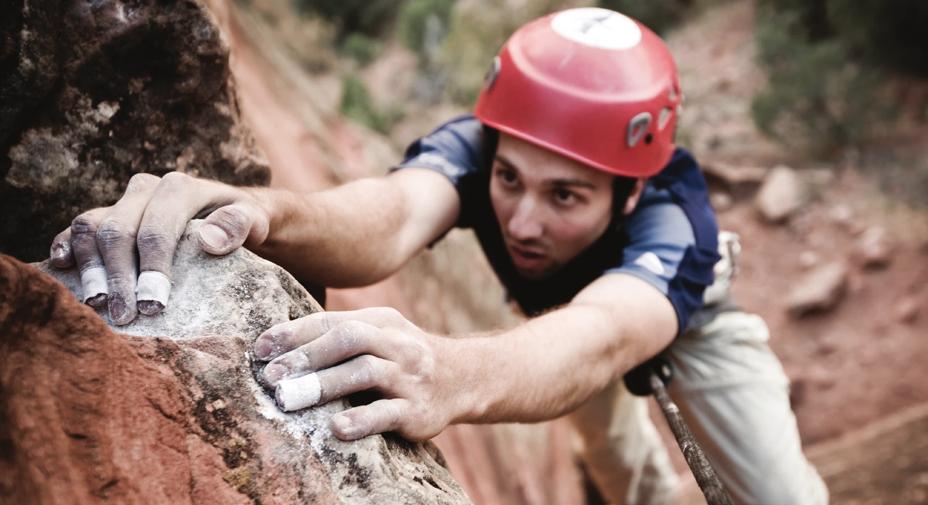 A climber topping out on a boulder problem, fingers taped, with a rope trailing below