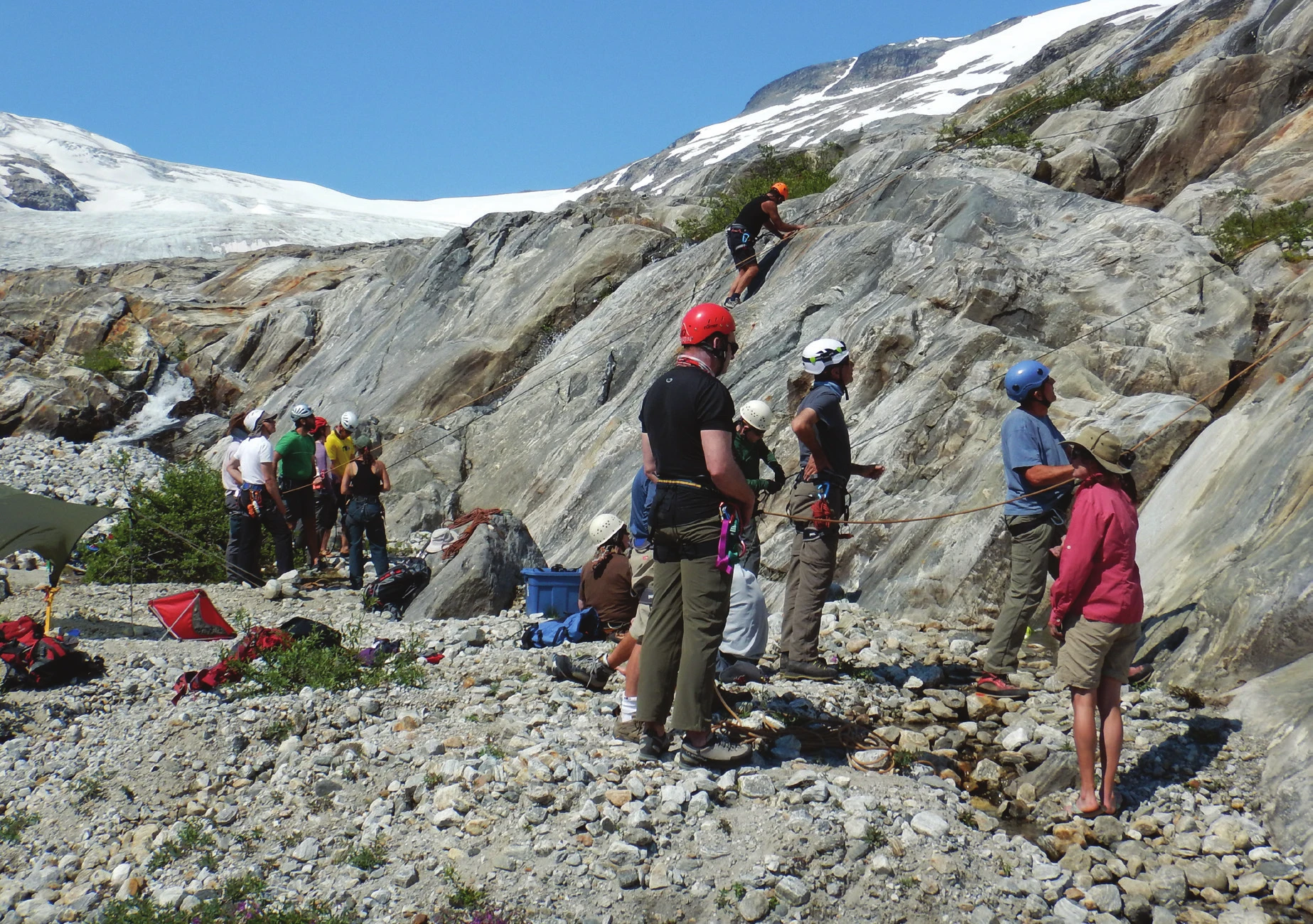 A top roping group gathered at the base of an alpine slab, with a climber high on the rock and a glacier behind